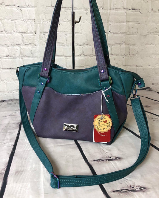 A classy handbag in two beautiful shades of dark teal and purple.  There is an adjustable, detachable crossbody strap.  Top handles for crook of the arm or shoulder.  Exterior has two large slip pockets. Interior has a zipper pocket and two slip pockets.  The two colors criss cross at the sides creating a beautiful unique pattern. Pretty lotus flower zipper pulls.  At time of this listing, 5 colors available. Message me with any questions.  12” W at the Base, 15” W at the Top, 9 h 6.25 deep