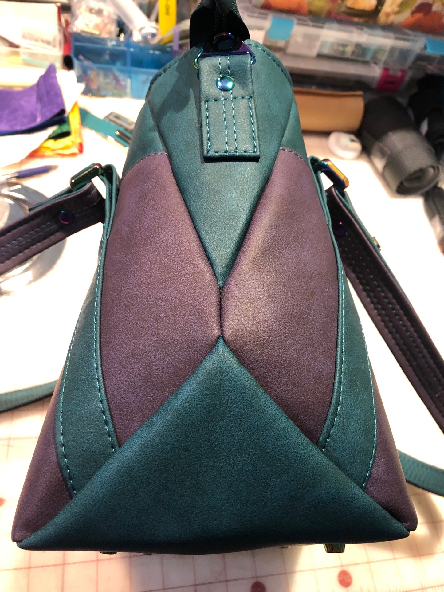 A classy handbag in two beautiful shades of dark teal and purple.  There is an adjustable, detachable crossbody strap.  Top handles for crook of the arm or shoulder.  Exterior has two large slip pockets. Interior has a zipper pocket and two slip pockets.  The two colors criss cross at the sides creating a beautiful unique pattern. Pretty lotus flower zipper pulls.  At time of this listing, 5 colors available. Message me with any questions.  12” W at the Base, 15” W at the Top, 9 h 6.25 deep