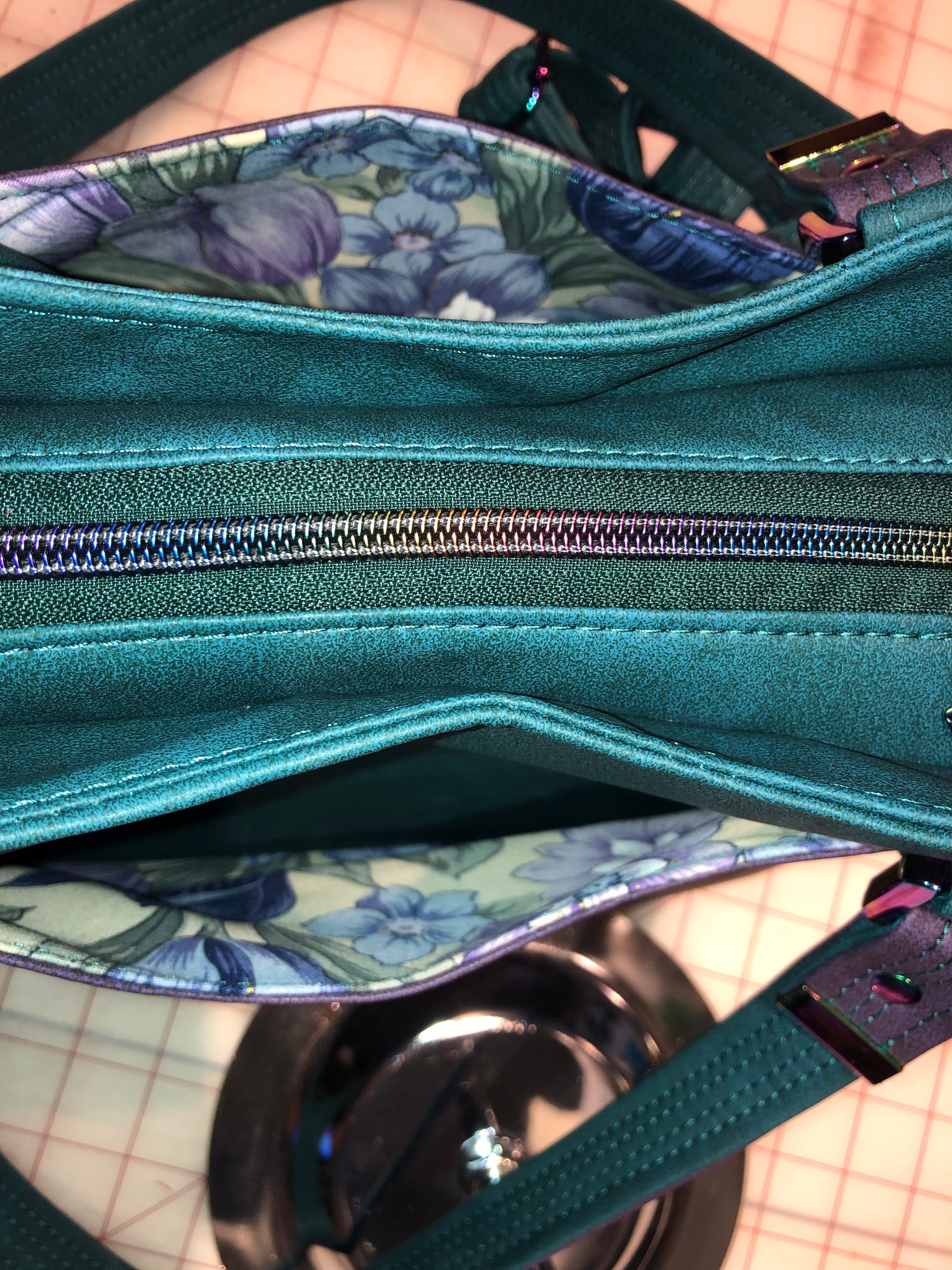 A classy handbag in two beautiful shades of dark teal and purple.  There is an adjustable, detachable crossbody strap.  Top handles for crook of the arm or shoulder.  Exterior has two large slip pockets. Interior has a zipper pocket and two slip pockets.  The two colors criss cross at the sides creating a beautiful unique pattern. Pretty lotus flower zipper pulls.  At time of this listing, 5 colors available. Message me with any questions.  12” W at the Base, 15” W at the Top, 9 h 6.25 deep